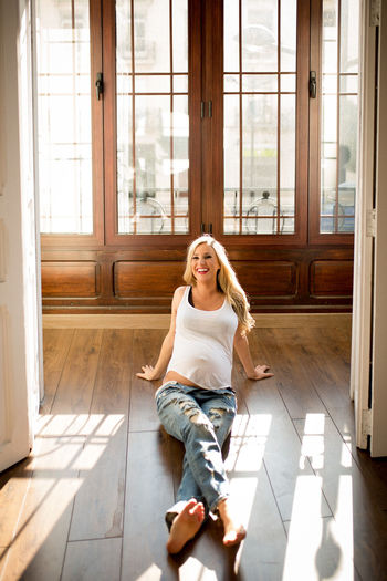 Happy pregnant female smiling and looking away while sitting on floor in spacious room on sunny day