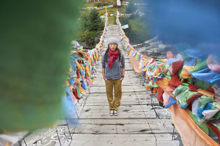 Young ethnic female tourist in warm clothing and hat looking at camera while crossing simple long narrow suspension wooden bridge decorated with bright multi colored pieces of fabric in daylight