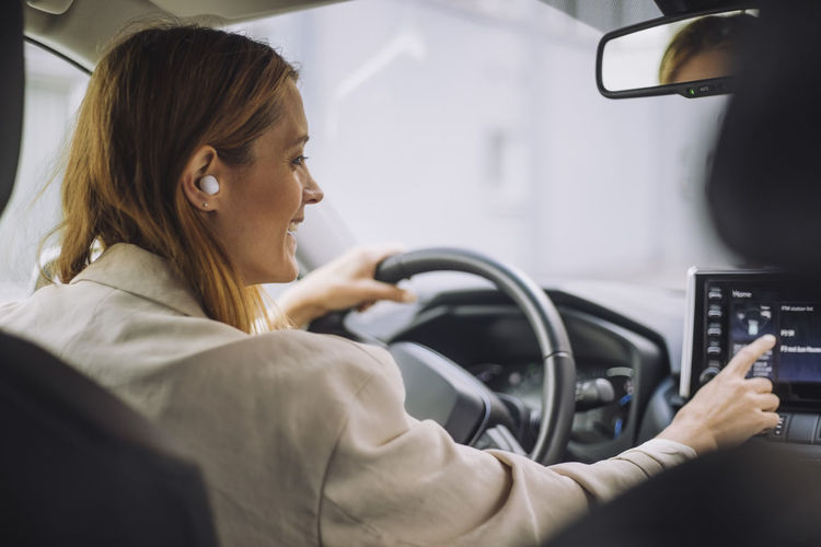 Smiling businesswoman wearing wireless in-ear headphones using dashboard control panel in car