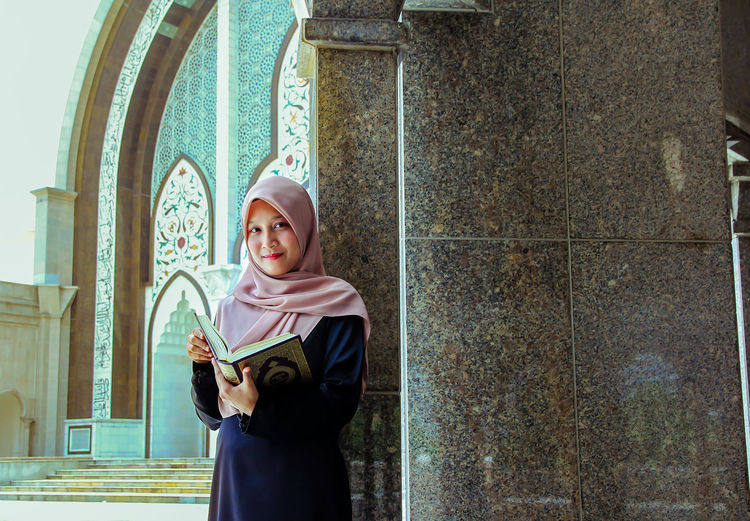 Portrait of young woman wearing hijab while reading holy book at mosque