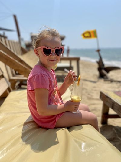 Portrait of girl wearing sunglasses with drink sitting at beach
