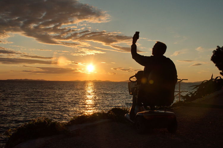 Disabled senior man sitting in wheelchair and using mobile phone during sunset on adriatic seashore