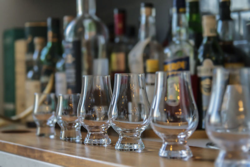 Old fashioned glasses lined up with many varieties of bottles in the background.