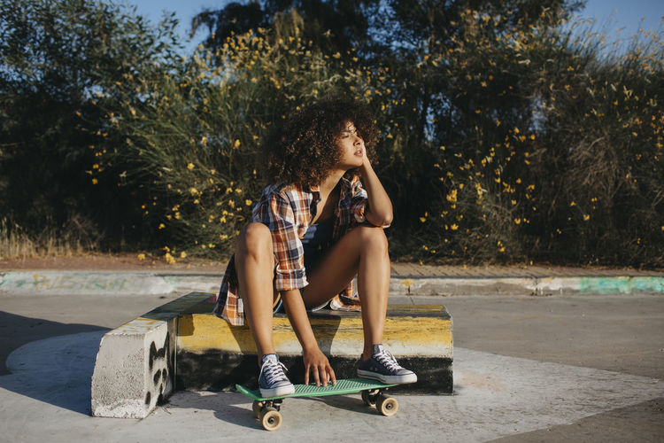 Calm black female with in trendy summer outfit and with afro hairstyle sitting on stone border with green penny board and looking away
