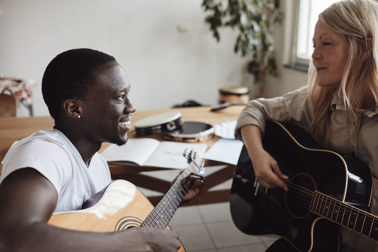 Smiling female mature tutor teaching guitar to young man in classroom