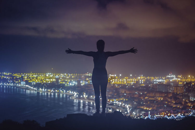 Rear view of woman with arms outstretched against cityscape at dusk