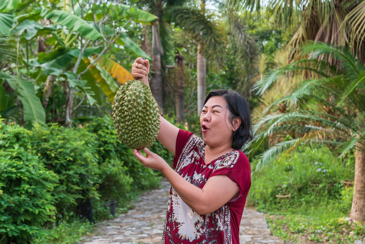 Shocked mature woman looking at durian while standing on footpath amidst plants