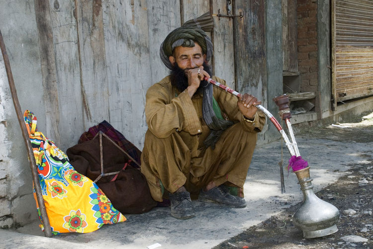 Full-length portrait of a serious young man sitting outdoors and smoking hukka.