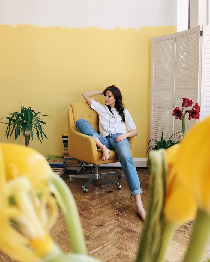YOUNG WOMAN SITTING ON CHAIR AT HOME AGAINST PLANTS