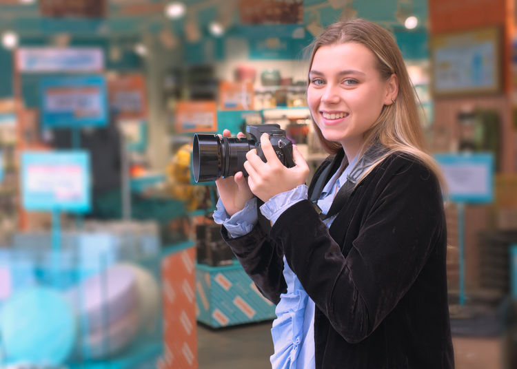 Portrait of smiling woman holding camera while standing at home