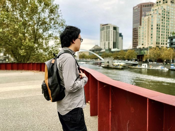 Young man standing on promenade and looking at river and buildings in the city.