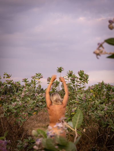 Rear view of sensuous naked woman standing by plants against sky