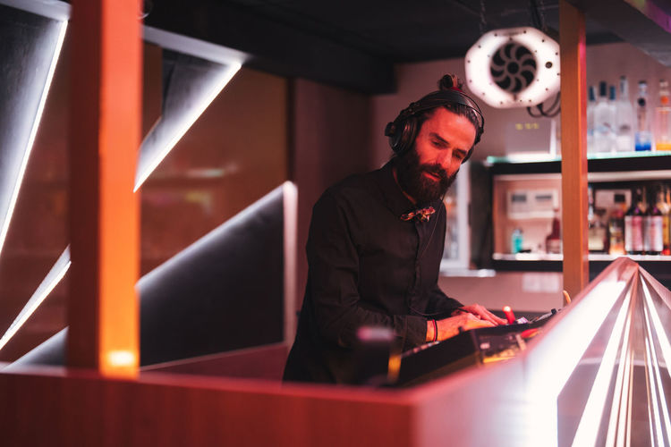 Concentrated young bearded male dj in headphones standing at illuminated counter in modern nightclub