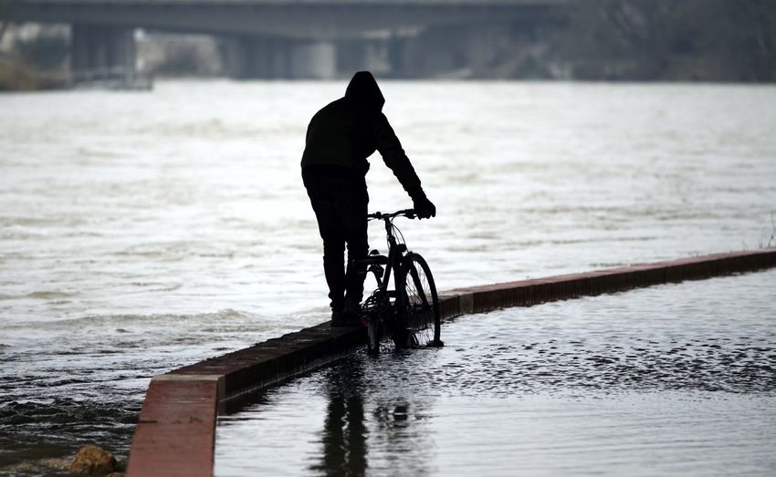 Rear view of silhouette man with bicycle walking on pier at riverbank
