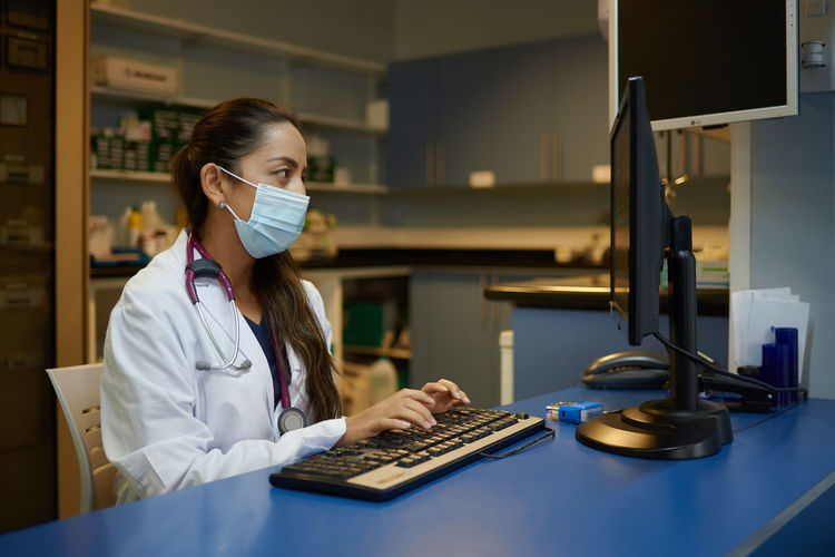 Side view of young female medic in white coat and protective mask typing on computer keyboard while working in cabinet of modern clinic