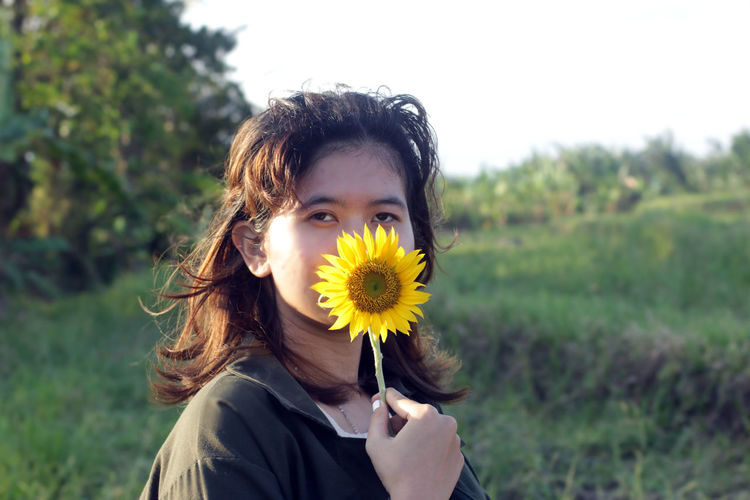 Portrait of a girl hiding or covering her half face with sunflower in the field. 