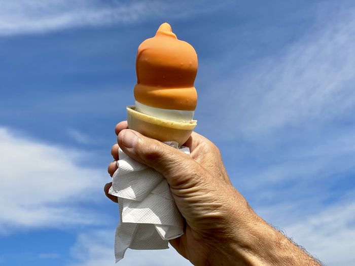 Cropped hand holding orange ice cream cone with napkin against sky