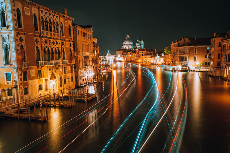 Light trails on river amidst illuminated buildings in city at night