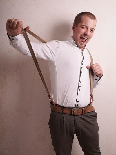 Portrait of man holding suspenders and standing against wall