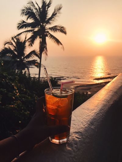Cropped image of hand holding drink against sea during sunset