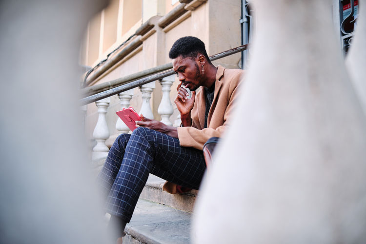 Modern thoughtful black african american man wearing stylish clothes and earring browsing on tablet while sitting in stone stairway on the street