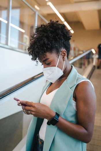 Side view of positive young african american female in casual outfit and protective mask using mobile phone while standing inside public building with escalator