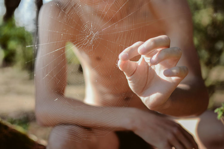 Midsection of shirtless man holding spider seen through web
