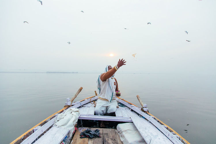 Varanasi, india - february, 2018: indian male in white clothes sitting in boat and throwing grains over river while feeding birds in early morning