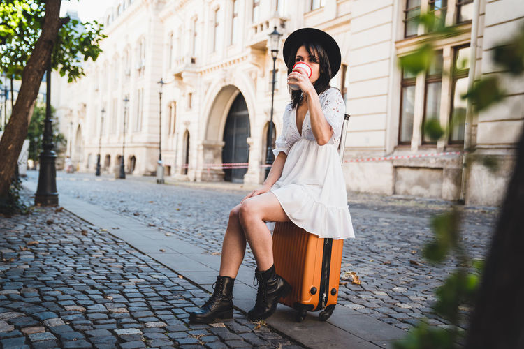 Full length of woman sitting on suitcase while drinking coffee