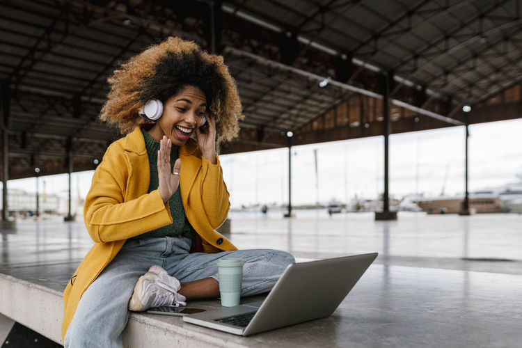Cheerful afro woman with in-ear headphones waving hand during video call through laptop in seating area