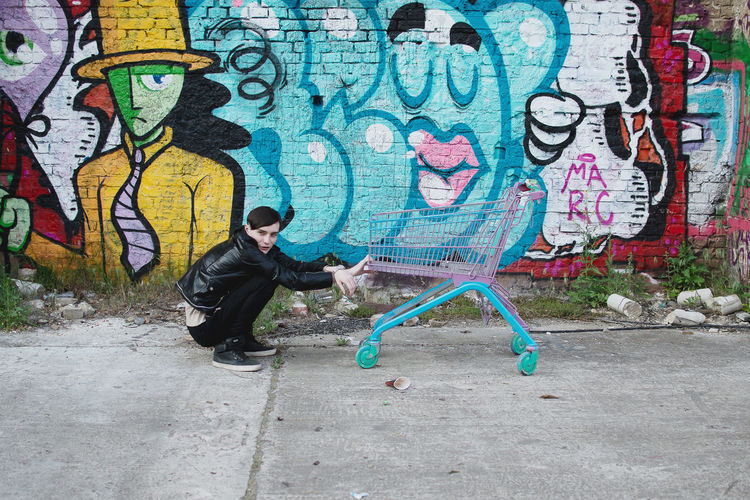 Side view of young man crouching by shopping cart against graffiti wall