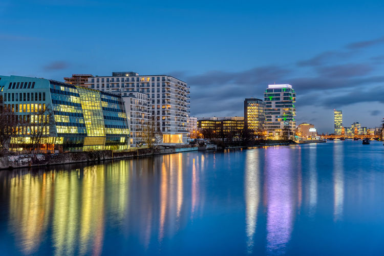 Illuminated buildings by river against blue sky