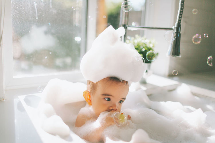 Cute baby boy taking bubble bath in kitchen sink at home