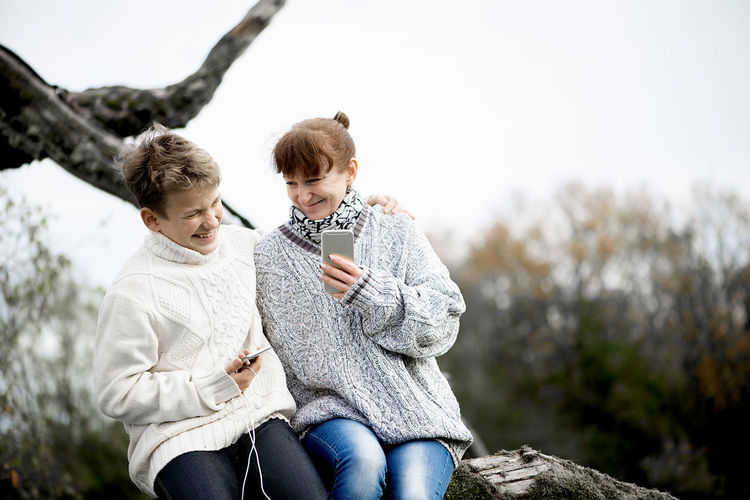 Happy mother and son teen sit together on a tree branch and use cell phones and headsets