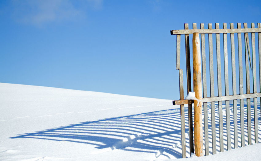 Shadow of fence on snow covered landscape against blue sky