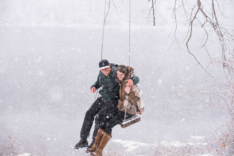 Couple laughing on swing over river during snowfall