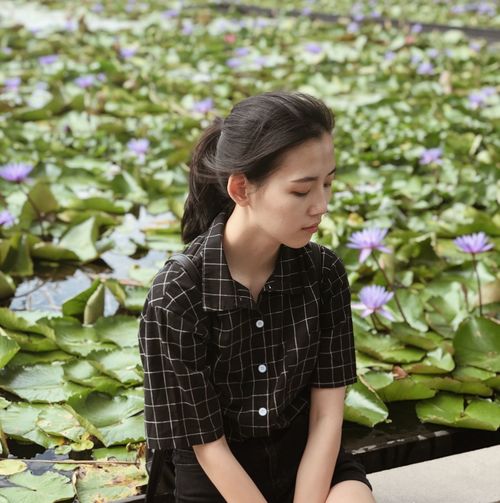 Sad young woman looking down while sitting by lotus plants in pond