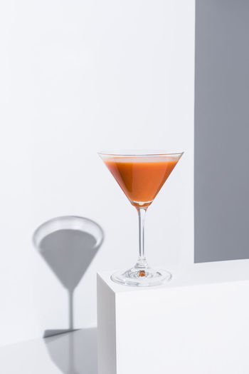 Cocktail glass with fresh red cold gazpacho soup placed on white stand on gray background in studio with bright sunlight