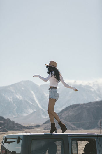 Full length of carefree young woman standing on off-road vehicle's roof at desert during sunny day