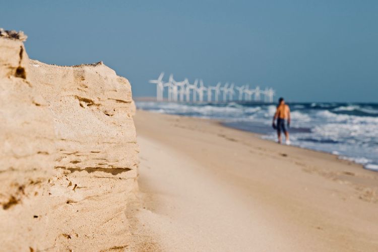 Rear view of shirtless man walking at beach against windmills