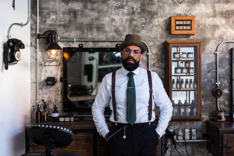 Confident ethnic bearded barber wearing classy outfit standing with hands in pockets in modern barbershop and looking at camera