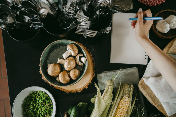 Hand of female chef writing recipe in diary by vegetables and cutlery on counter