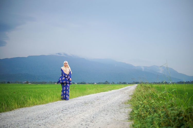 Woman standing on road amidst field against sky