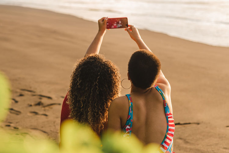 Back view of unrecognizable young ethnic women taking selfie on mobile phone while enjoying summer vacation together on sandy seashore in costa rica
