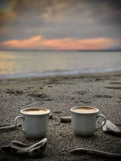 Close-up of coffee cup on sea shore against sunset sky