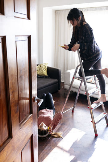 Young woman on ladder taking picture of friend lying down at home