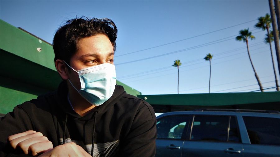 Close-up of young man wearing flu mask looking away standing against sky