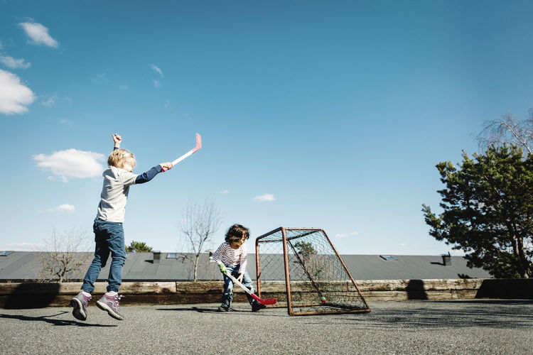Excited girl playing hockey with boy at yard against blue sky