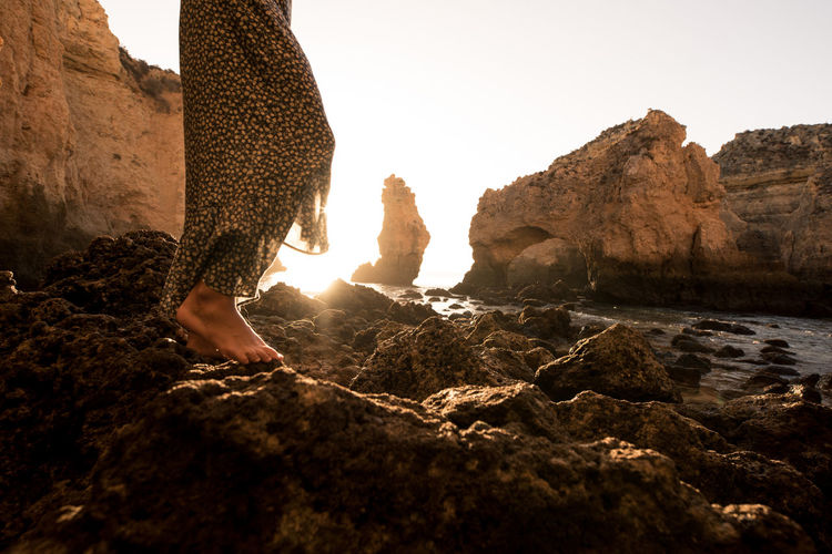 Back view of cropped anonymous female standing barefoot in rocks near sea on sunny day in algar seco caves in algarve, portugal