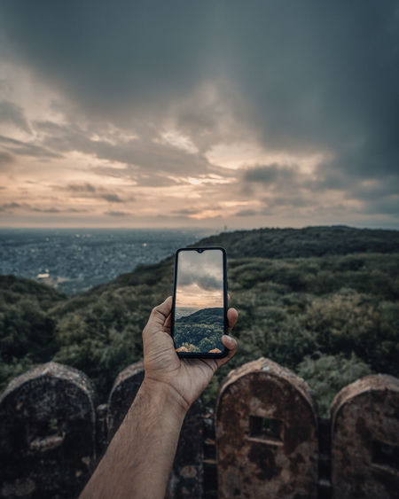 Man holding smart phone by sea against sky during sunset